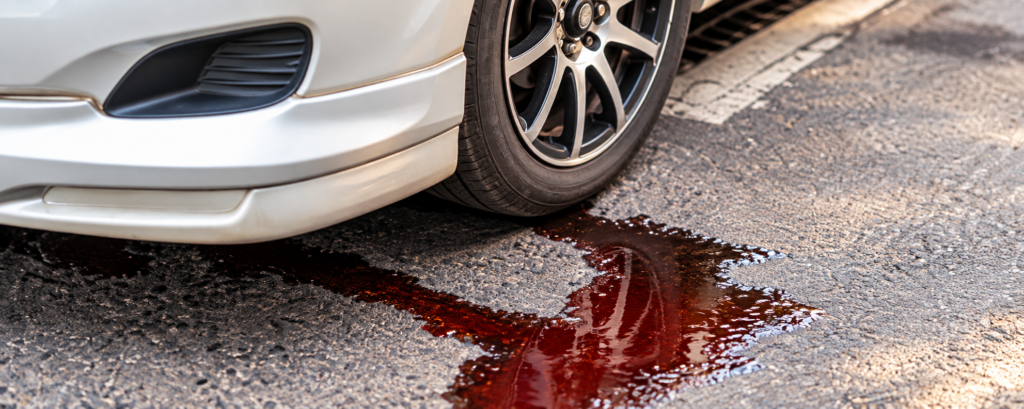 A puddle of dark red fluid leaking onto the pavement from underneath the front of a silver car, indicating a potential transmission leak.