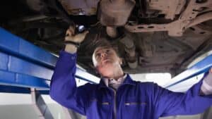 A mechanic in a blue jumpsuit inspects the underside of a car using a flashlight while the vehicle is lifted on a hydraulic car lift, beginning a stress-free diagnostic process for potential transmission repair.