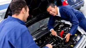 Two mechanics in blue coveralls work on a car engine together in a South Salt Lake, UT garage, both smiling as they tackle driveline repair under the open hood.