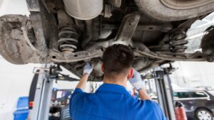 An expert mechanic in blue overalls inspects the underside of a car lifted on a hydraulic lift in an auto repair shop, demonstrating the high standards of auto repair available in White City UT.