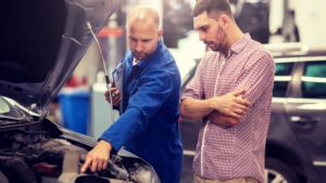 An expert mechanic in blue overalls explains engine issues to a man in a checked shirt, both standing by a car with its hood open in an auto repair garage in White City, UT.
