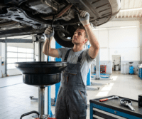 Mechanic in overalls works under a raised car in a brightly lit auto repair shop, holding a tool by an oil drain pan during professional oil changes to help prevent engine wear and ensure proper car maintenance.