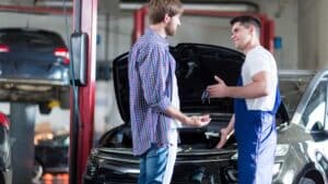 A mechanic in blue overalls discusses driveline repair with a man in casual clothes next to a car with its hood open inside a Riverton auto repair shop.
