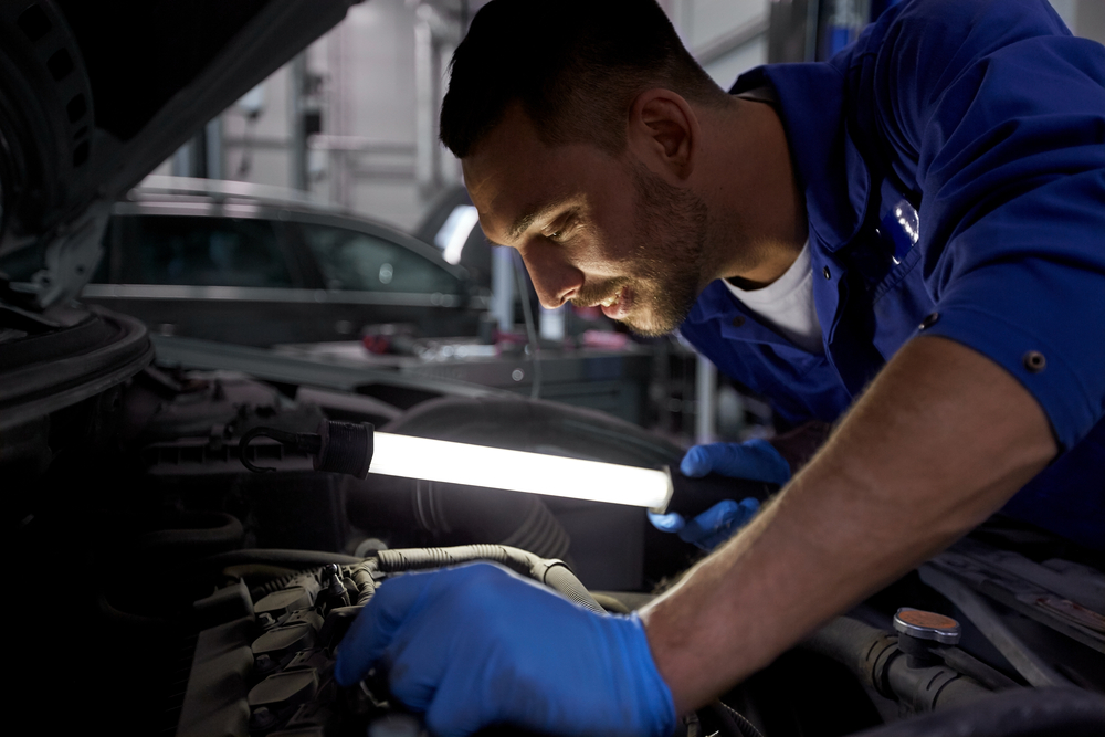 A mechanic wearing blue gloves inspects a car engine under bright lighting in an automotive workshop.